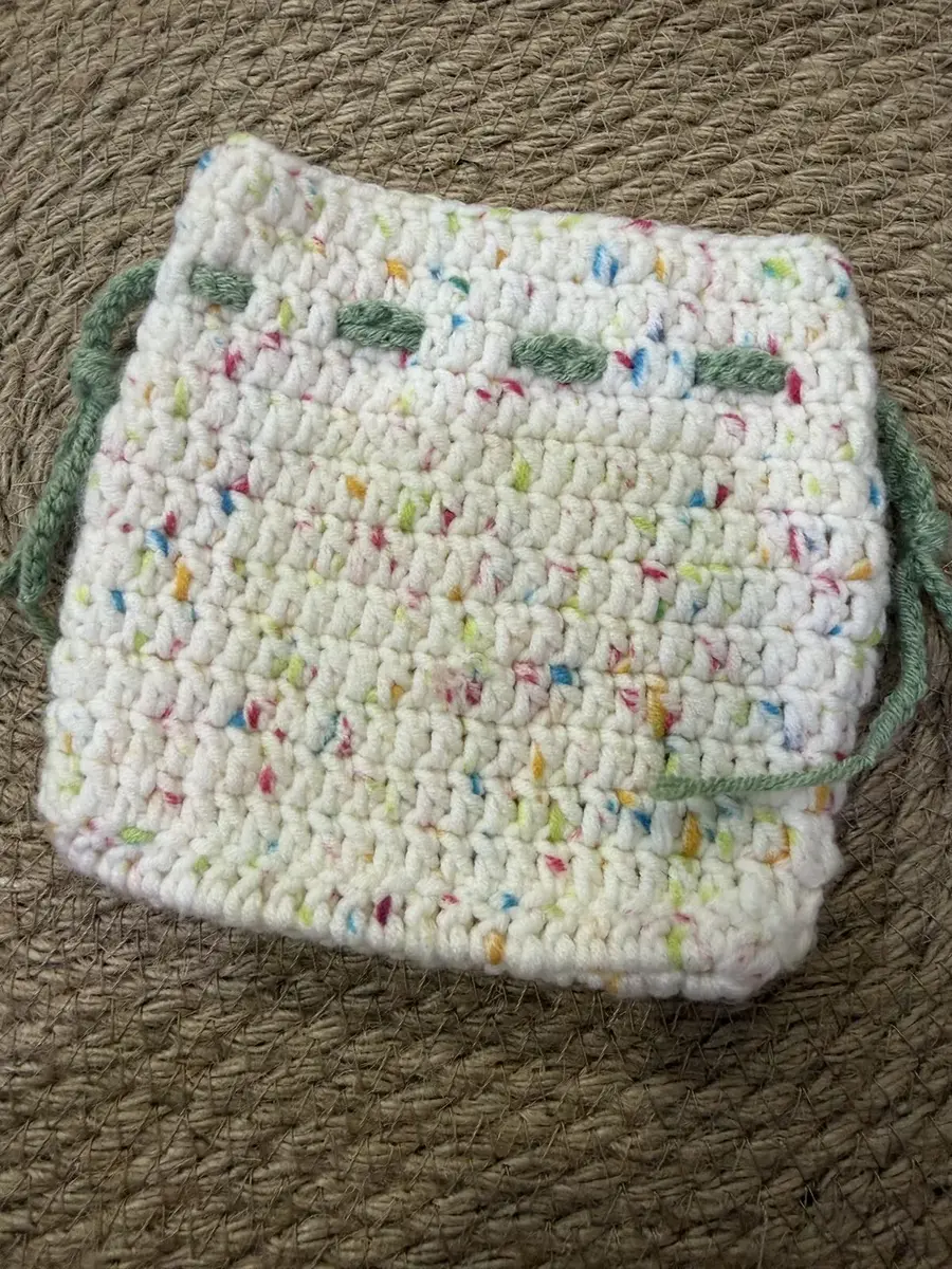A crochet drawstring pouch in cream with rainbow confetti and a green tie.